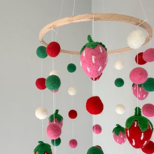 May include: A felt baby mobile with a wooden ring and hanging felt balls in red, green, white, and pink. The mobile features felt strawberries in pink and red.