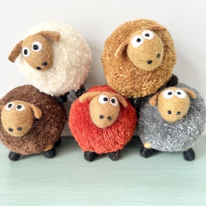 May include: A collection of handmade sheep figurines in various colors. The sheep have fluffy wool bodies in white, brown, red, gray, and tan. Each sheep has a tan face with black eyes and ears, and black feet.