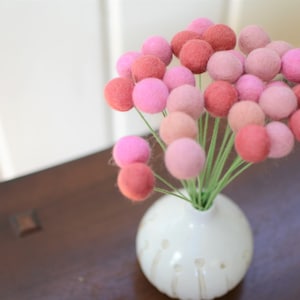 May include: A bouquet of pink and red felt ball flowers in a white vase with a textured surface.
