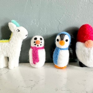May include: Four felt animal ornaments: a white unicorn with a yellow and blue mane, a white snowman with a pink scarf, a white penguin with a blue scarf, and a white gnome with a red hat.