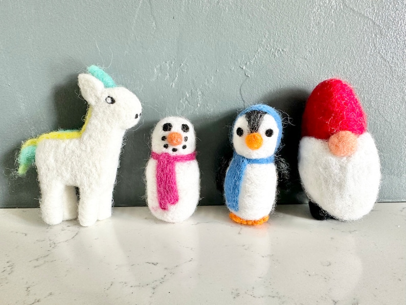 May include: Four felt animal ornaments: a white unicorn with a yellow and blue mane, a white snowman with a pink scarf, a white penguin with a blue scarf, and a white gnome with a red hat.