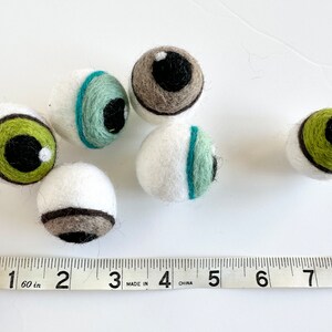 May include: Six felt eyeballs in various colors, including green, brown, and blue, with black pupils. The eyeballs are approximately 1 inch in diameter and are arranged on a white surface with a measuring tape.