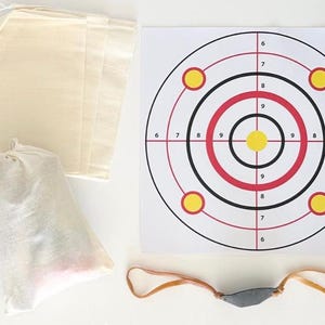 May include: A set of white fabric bags, a slingshot with a gray pouch, and a paper target with red and black rings and yellow circles. The target has numbers 6, 7, 8, and 9 printed on it.