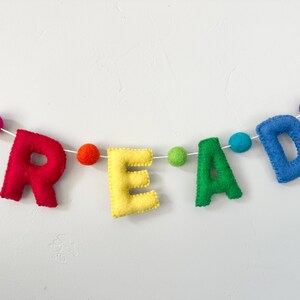 Puede incluir: Una pancarta de fieltro con la palabra "READ" en colores arco&iacute;ris. Las letras est&aacute;n enhebradas con hilo blanco y peque&ntilde;os pompones en colores coordinados.