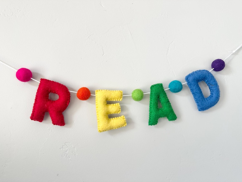 Puede incluir: Una pancarta de fieltro con la palabra "READ" en colores arco&iacute;ris. Las letras est&aacute;n enhebradas con hilo blanco y peque&ntilde;os pompones en colores coordinados.