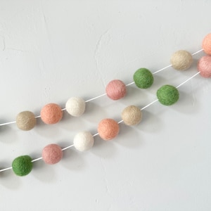 May include: A garland of felt balls in shades of pink, green, and beige. The balls are strung on a white string and are arranged in a diagonal pattern.