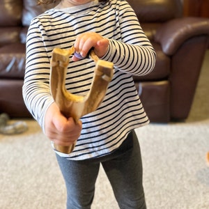 K&ouml;nnte beinhalten: Eine Holzschleuder mit hellbraunem Finish wird von einer Person gehalten, die ein gestreiftes Langarmshirt und graue Leggings tr&auml;gt. Die Schleuder wird in Schussposition gehalten, bereit, ein Projektil abzufeuern.