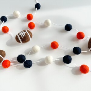 Puede incluir: Una guirnalda con bolas de fieltro blancas, naranjas y azul marino y balones de f&uacute;tbol de fieltro marrones.