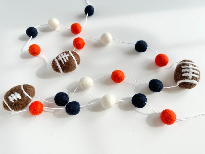 Puede incluir: Una guirnalda con bolas de fieltro blancas, naranjas y azul marino y balones de f&uacute;tbol de fieltro marrones.