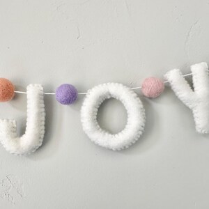 May include: A decorative garland spelling the word JOY in white felt letters, interspersed with small felt balls in pastel shades of peach, purple, pink, and mint green, all strung together on a thin white cord for hanging display.