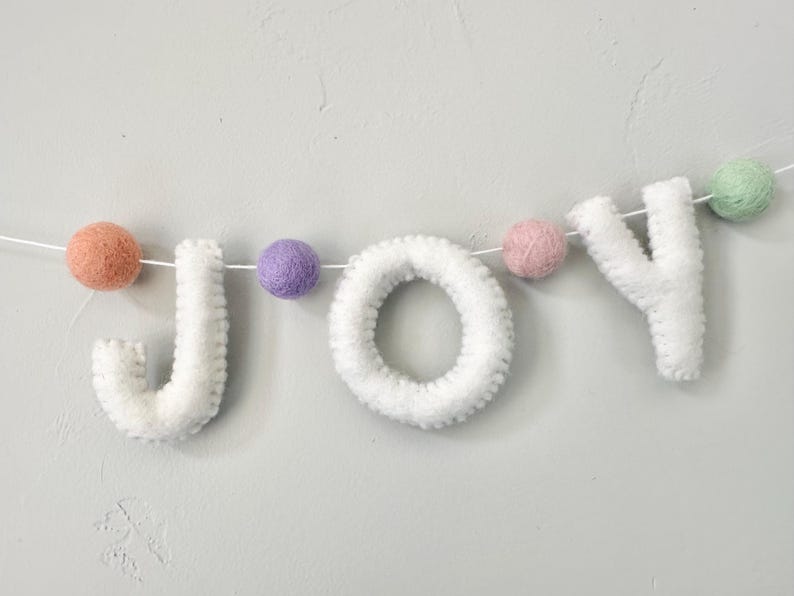 May include: A decorative garland spelling the word JOY in white felt letters, interspersed with small felt balls in pastel shades of peach, purple, pink, and mint green, all strung together on a thin white cord for hanging display.