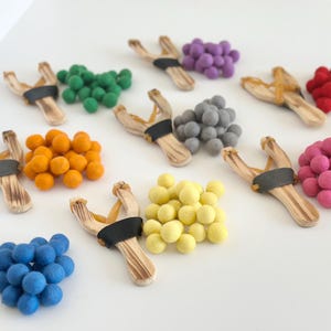 May include: A collection of colorful slingshots with felt ball ammunition. The slingshots are made of light-colored wood with black rubber bands. The felt balls are in various colors, including green, orange, purple, gray, red, pink, yellow, and blue.