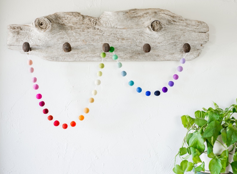 May include: Three colorful felt ball garlands hanging from a rustic wooden branch. The garlands are made of different shades of pink, orange, yellow, green, blue, and purple.
