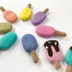 May include: A collection of colorful felt ice cream popsicles in various pastel shades, including pink, blue, green, and yellow. Each popsicle has a brown stick and some have chocolate drizzle details. These handmade felt toys are perfect for children.