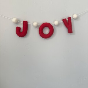 May include: A decorative garland featuring the word JOY spelled out in red felt letters, interspersed with small white felt balls. The letters and balls are strung together on a thin white cord, creating a festive banner for seasonal display.