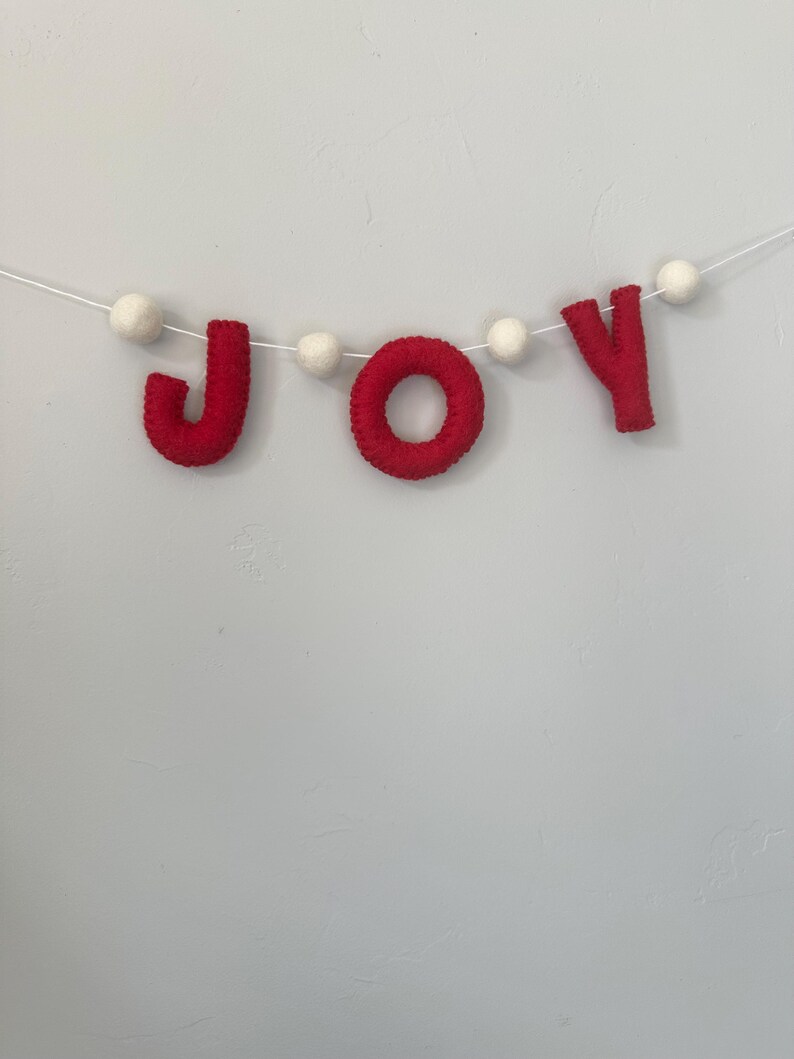 May include: A decorative garland featuring the word JOY spelled out in red felt letters, interspersed with small white felt balls. The letters and balls are strung together on a thin white cord, creating a festive banner for seasonal display.