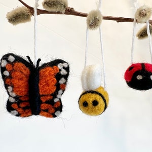 May include: Three felt ornaments hanging from a branch. The ornaments are a butterfly with orange and black wings, a bee with yellow and black stripes, and a ladybug with a red shell and black spots.