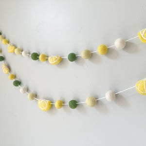 May include: A garland made of felt balls in white, yellow, and green. The yellow balls are shaped like lemon slices. The garland is strung on white thread.