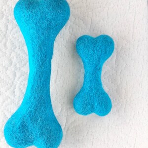 May include: Two blue felt dog bones, one larger than the other, on a white textured surface.