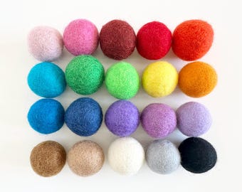 May include: A collection of twenty-one colorful felt balls in various shades of pink, red, orange, yellow, green, blue, purple, brown, gray, and black. These decorative balls are arranged in rows on a white surface.