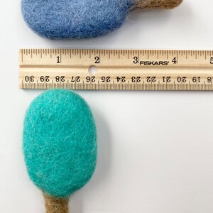 May include: Two felted wool ornaments, one blue and one teal, each with a brown stem. A wooden ruler shows the ornaments are approximately 3 inches long. The ornaments are handmade and have a soft, textured appearance.