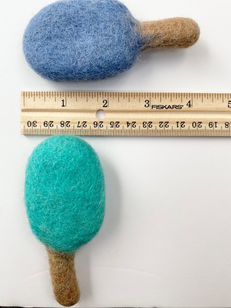 May include: Two felted wool ornaments, one blue and one teal, each with a brown stem. A wooden ruler shows the ornaments are approximately 3 inches long. The ornaments are handmade and have a soft, textured appearance.