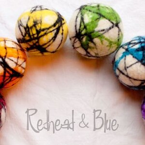 May include: Seven colorful felt balls arranged in an arc. Each ball is a different color, including red, orange, yellow, green, blue, and purple, with a white base and black web-like design. The text "Redhead & Blue" is at the bottom.