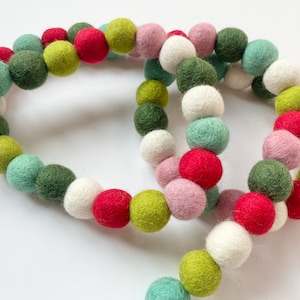 Puede incluir: Una guirnalda de bolas de fieltro en tonos de verde, rojo, blanco y rosa. Las bolas están enhebradas para crear una decoración festiva.