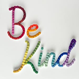 May include: Wall decor spelling the words Be Kind, constructed from small, spherical felt balls in a vibrant rainbow gradient. The letters are arranged in a cursive script, transitioning through shades of red, orange, yellow, green, blue, and purple.