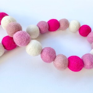 May include: A garland of felt balls in pink, white, and light pink colors. The garland is made of soft, fluffy felt balls that are strung together on a string.