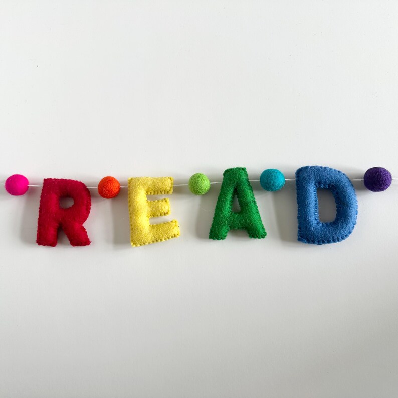 Puede incluir: Una pancarta de fieltro con la palabra "READ" en colores arco&iacute;ris. Las letras est&aacute;n enhebradas con hilo blanco y separadas por pompones de colores.