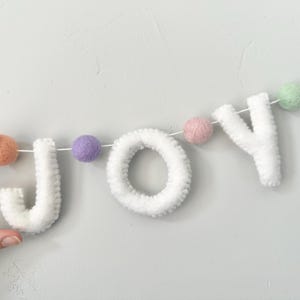 May include: A garland spelling the word JOY in white felt letters, interspersed with small, round, multi-colored felt balls in shades of peach, purple, pink, and light green, all strung together on a thin white cord for hanging display.