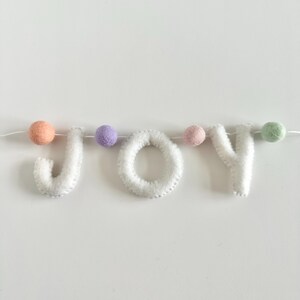 May include: A decorative garland spelling the word JOY in white felt letters, interspersed with small felt balls in peach, lavender, light pink, and mint green colors, all strung together on a thin white cord for hanging display.