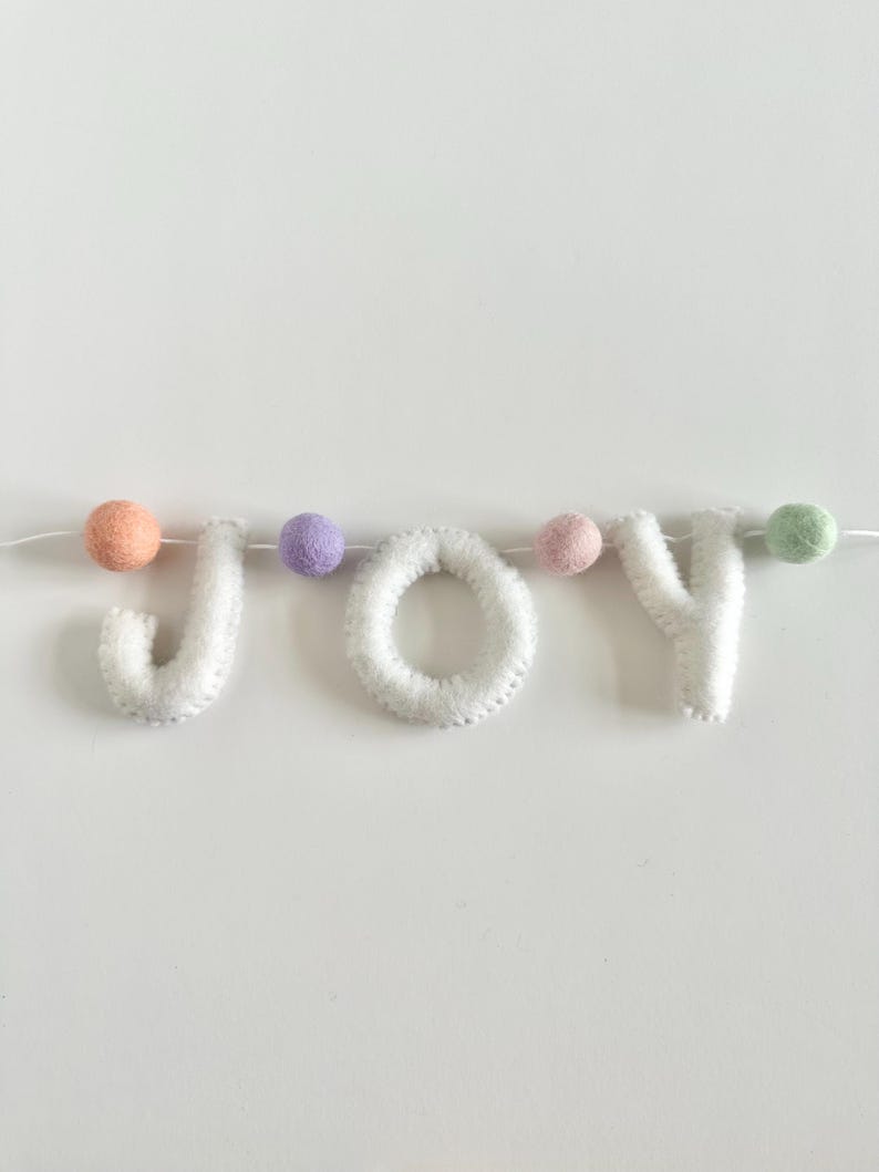 May include: A decorative garland spelling the word JOY in white felt letters, interspersed with small felt balls in peach, lavender, light pink, and mint green colors, all strung together on a thin white cord for hanging display.