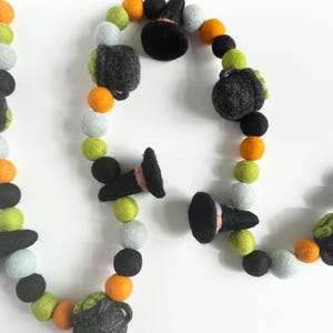 May include: A Halloween garland made of felt balls in orange, green, gray, and black. The garland features witch hats and cauldrons.