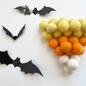 May include: A Halloween garland made of felt balls in yellow, orange, and white. The garland is shaped like a candy corn and is decorated with three black paper bats.