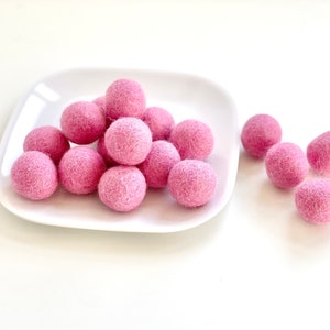 May include: A pile of pink felt balls on a white square plate. There are also several pink felt balls scattered on a white surface.