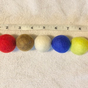 May include: Eight felt balls in different colors: yellow, red, brown, white, blue, green, orange. The balls are lined up in a row on a white surface with a measuring tape behind them.