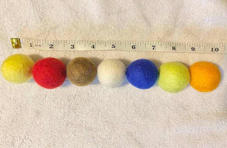 May include: Eight felt balls in different colors: yellow, red, brown, white, blue, green, orange. The balls are lined up in a row on a white surface with a measuring tape behind them.
