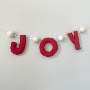 May include: A festive garland featuring the word JOY spelled out in red felt letters, interspersed with small white felt balls strung along a thin white cord. The letters J, O, and Y are crafted from soft red material, creating a clear, bold display.