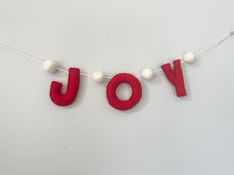 May include: A festive garland featuring the word JOY spelled out in red felt letters, interspersed with small white felt balls strung along a thin white cord. The letters J, O, and Y are crafted from soft red material, creating a clear, bold display.