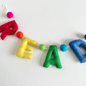 Puede incluir: Una pancarta de fieltro que deletrea la palabra "READ" en colores arco&iacute;ris con pompones entre cada letra.