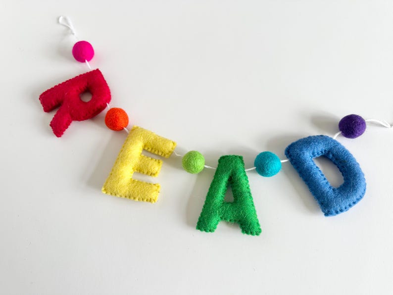 Puede incluir: Una pancarta de fieltro que deletrea la palabra "READ" en colores arco&iacute;ris con pompones entre cada letra.