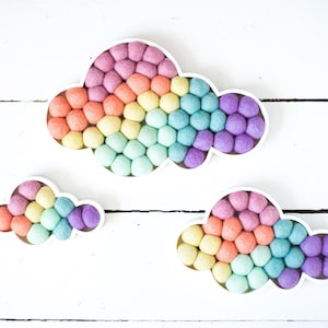Puede incluir: Tres decoraciones de pared con forma de nube con un marco blanco relleno de pequeñas bolas de fieltro redondas en un degradado de colores arcoíris que incluye rosa, naranja, amarillo, verde, azul y morado, con un patrón texturizado.