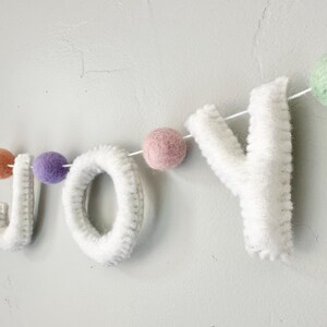May include: A felt garland spelling the word JOY in white, stitched letters. The letters are interspersed with small, round felt balls in muted shades of orange, purple, pink, and light green, all strung together on a thin white cord.