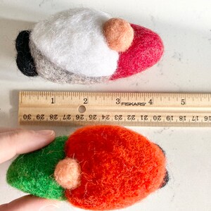 May include: Two felt gnome ornaments, one red and white, the other green and orange, with a brown felt beard and a small peach-colored felt nose. The ornaments are approximately 2 inches long and are measured against a ruler.