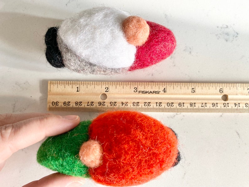 May include: Two felt gnome ornaments, one red and white, the other green and orange, with a brown felt beard and a small peach-colored felt nose. The ornaments are approximately 2 inches long and are measured against a ruler.