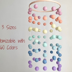 May include: A rainbow-colored felt ball mobile with a wooden ring. The mobile features a gradient of colors, starting with pink and transitioning to purple. The text "3 Sizes Customizable with 60 Colors" is visible in the image.