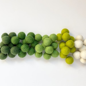 May include: A garland of felt balls in shades of green and white. The balls are arranged in a gradient from dark green to light green to white.