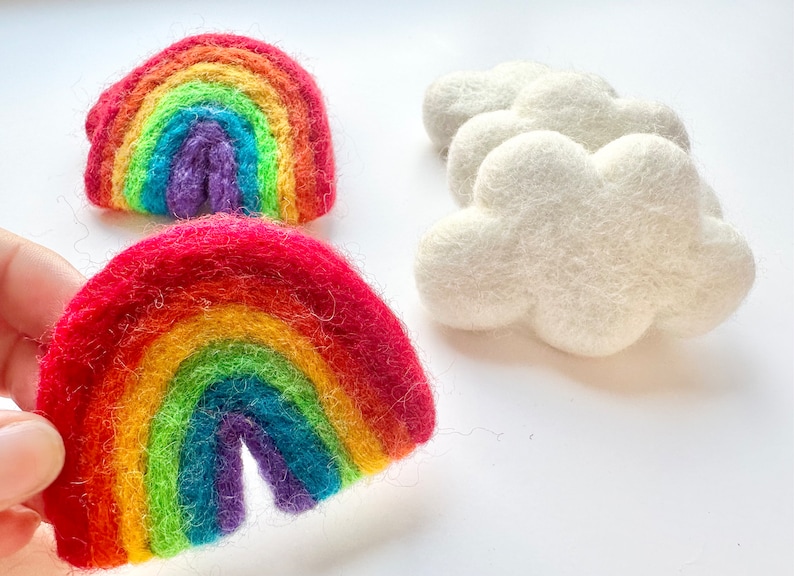 May include: Two felt rainbows, one red, orange, yellow, green, blue, indigo, and violet, and the other a similar colour scheme but with a lighter red, orange, and yellow.  The rainbows are handmade and have a fuzzy texture.  There are three white felt clouds in the background.